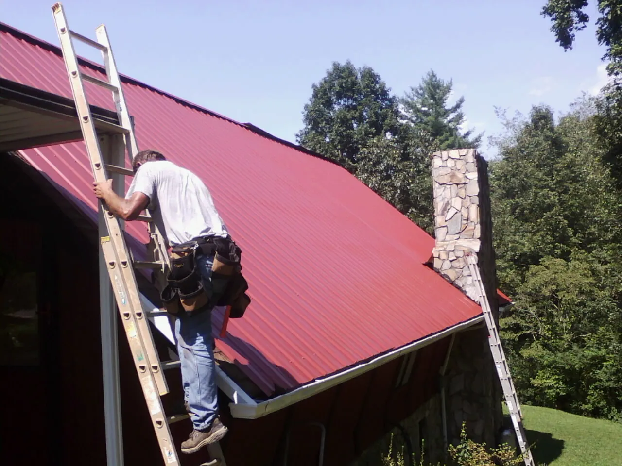 Roofer on a ladder beside a red metal roof and stone chimney — Soffit & Fascia Repair by Roofing Brothers in Mount Gretna